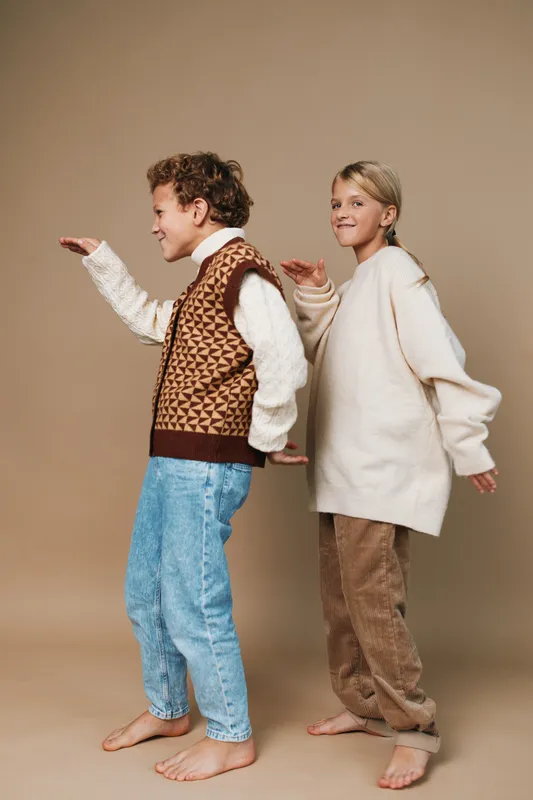 Two joyful figures pose in a studio, back to back, arms extended as if dancing. Casual sweaters and relaxed denim create a warm, friendly mood with playful movement.