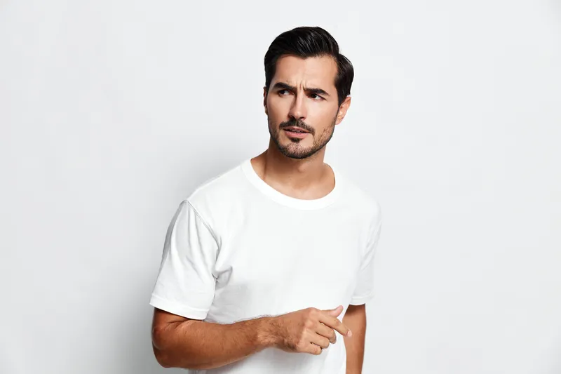 Handsome man with a curious expression wearing a white t-shirt, looking to the side while pointing, against a plain light background. Lifestyle concept.