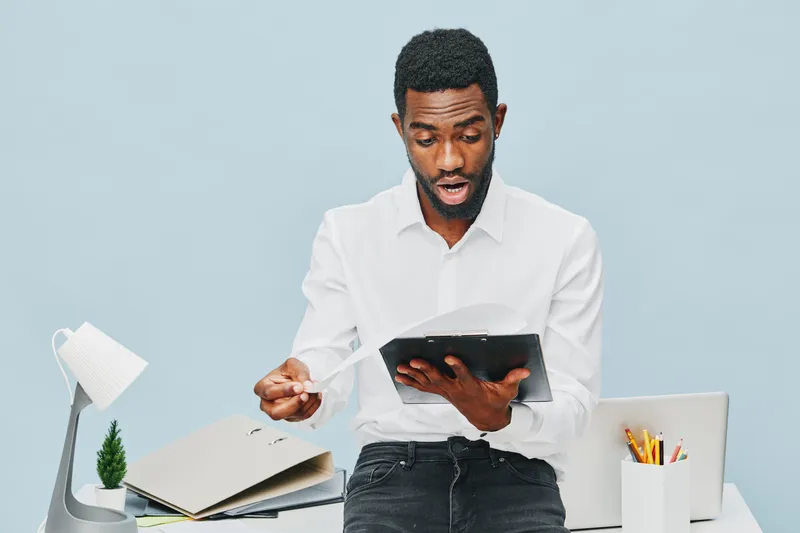 Professional young African American man in white shirt holding a tablet and reading, sitting at a desk with office supplies, isolated on light blue background. Business, technology, communication, wor
