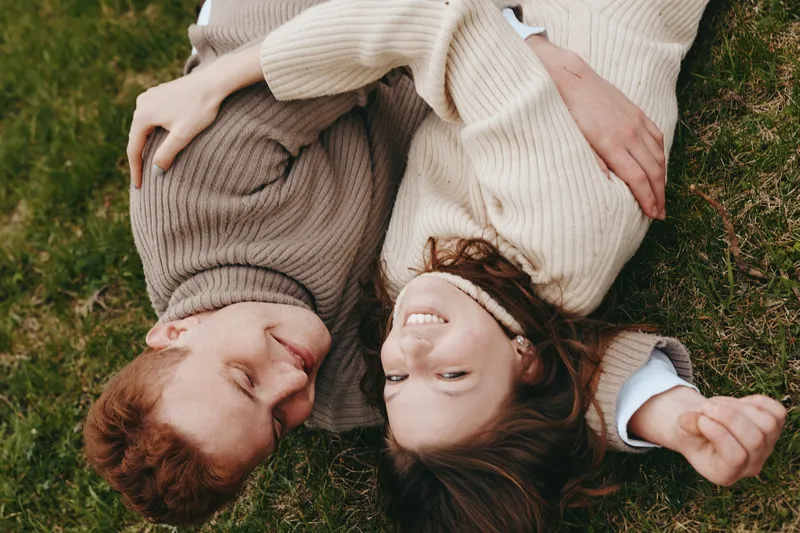 Young couple with red and brunette hair lies on green grass wearing cozy sweaters and shares a joyful moment outdoors in a natural lifestyle setting with relaxed smiles.