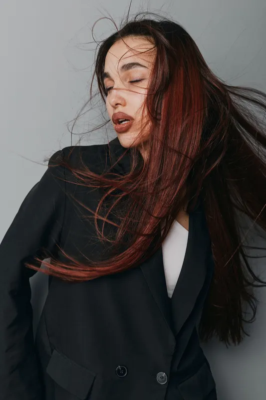Woman with long hair wearing black blazer posing with eyes closed and hair flowing in studio against gray background. Fashion model expressing emotion with natural makeup and stylish outfit.