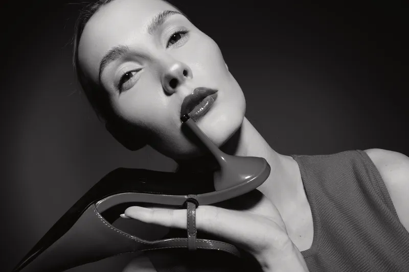 Close-up portrait of a caucasian woman with light skin holding a high heel shoe near her lips in black and white. Concept for fashion, elegance, and beauty in studio lighting.