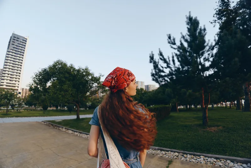 Lifestyle woman with long hair in modern boho-western denim street style walking outdoors, wearing a red bandana and casual bag, film color tones, urban park background.