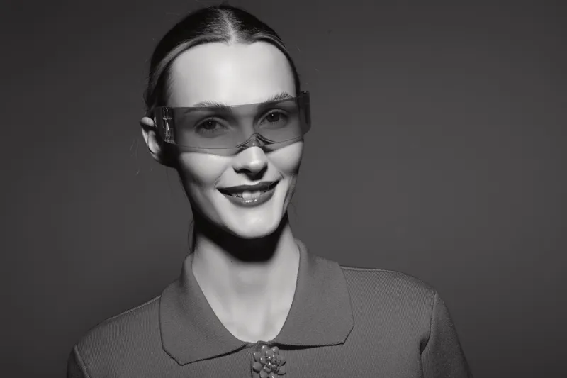 Smiling white woman with pale skin and slicked hair wearing large transparent sunglasses and a textured shirt in monochrome studio lighting. Concept of fashion, eyewear, and style.