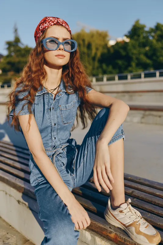 Lifestyle woman in modern boho-western denim street style wearing blue sunglasses and a red headband, sitting outdoors with a film color effect, casual urban outfit.