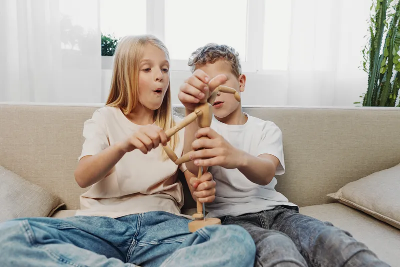 Two children sitting on a couch play with a wooden artist mannequin, showing curiosity and engagement in a bright living room. Concept of creativity, childhood, and learning through play.