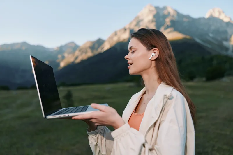 A focused woman stands outdoors in a scenic landscape, holding a laptop and looking toward the horizon. The serene setting suggests remote work, digital creativity, and nature inspired productivity.