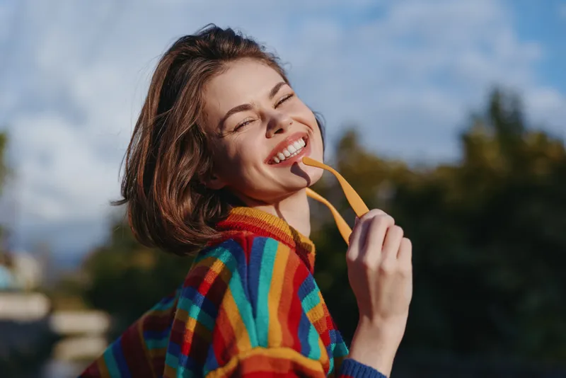 Young woman smiling in her mid twenties outdoors, wearing a colorful striped sweater and holding sunglasses strap, casual travel lifestyle portrait with sunny, joyful and carefree vibe.