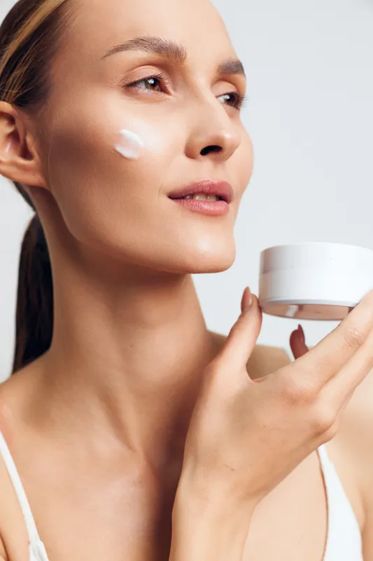 Young woman applying cream on face with smooth skin holding white jar, skincare routine, natural light, minimal makeup, clean background, beauty concept