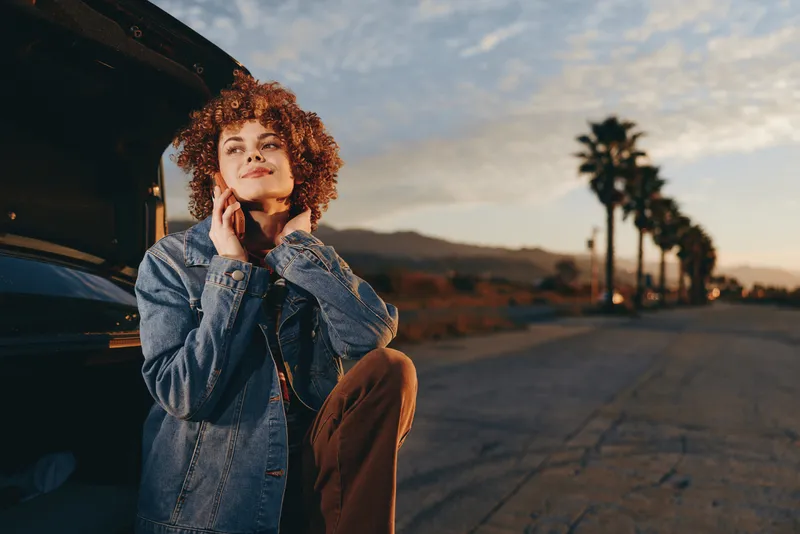 Woman with curly red hair stands outdoors on a street at sunset, holding a phone to her ear, wearing a denim jacket, casual lifestyle, warm light, city travel feeling, relaxed mood