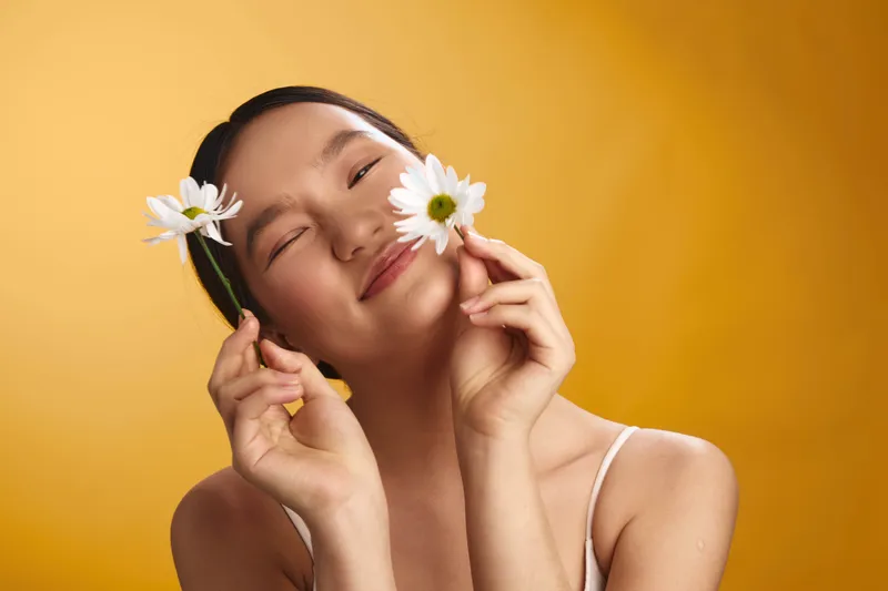 asian woman in a bright setting smiles softly while holding daisies near her cheeks, creating a joyful, fresh mood that radiates beauty and confidence in a playful springtime atmosphere.