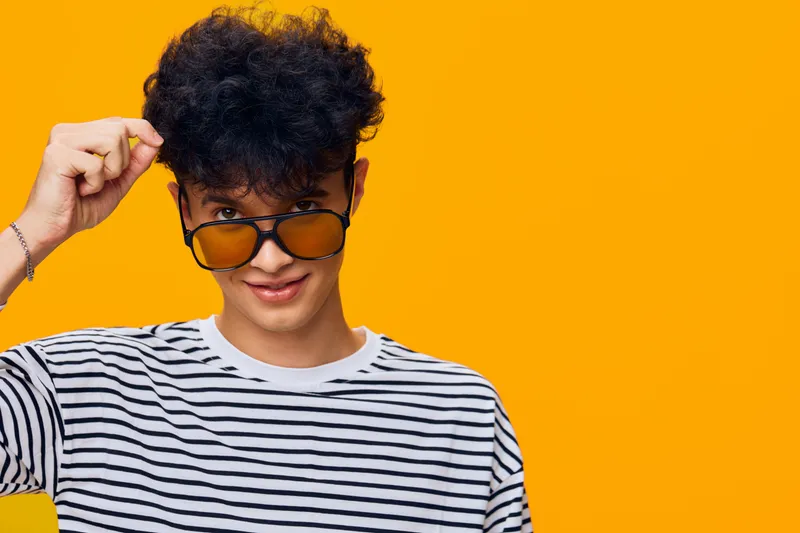 Young man wearing striped shirt and sunglasses with curly hair posing against vibrant yellow background, showing playful and confident expression.