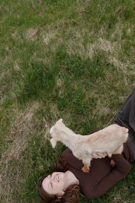 Young woman lies on grass smiling happily while holding a small white baby goat in a casual brown sweater in a natural outdoor lifestyle setting.