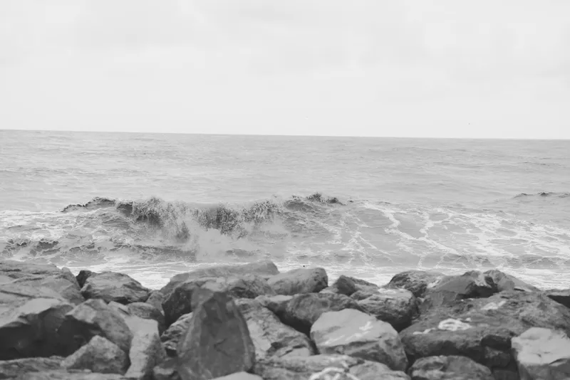 Monochrome scene with rocks along a calm shoreline as waves crash against the coast, texture of stone and foam under an overcast sky