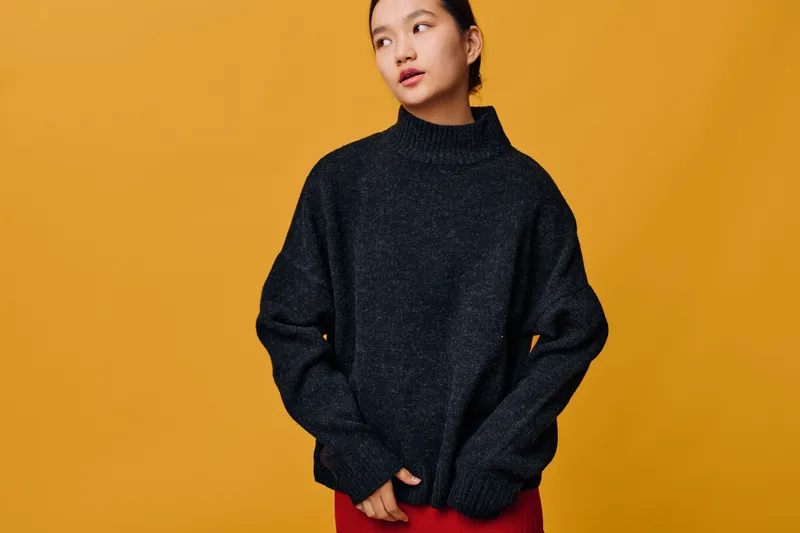 Young woman wearing oversized black sweater and red skirt posing on yellow background looking aside with thoughtful expression in studio for fashion portrait concept.