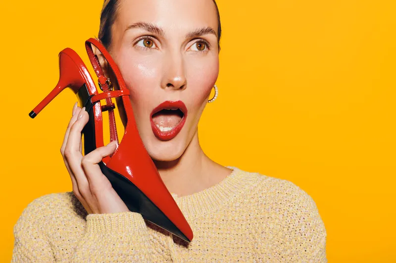Surprised woman holds red high heel shoe near ear as if it is a phone. Bright yellow background and casual sweater create playful and creative concept of fashion and communication.