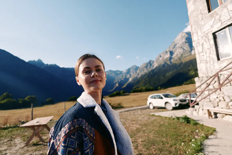 Woman portrait in the mountains beside a stone cottage and parked car, wearing a patterned jacket with shearling trim, smiling in rural countryside under clear blue sky.