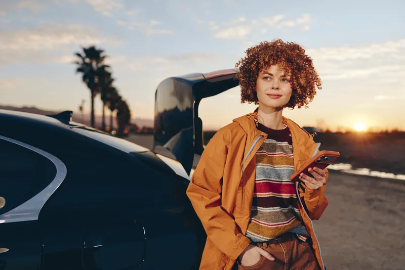 Woman lifestyle travel scene, standing by open car door on road at sunset, orange jacket and striped sweater, holding travel papers, relaxed portrait, evening light soft