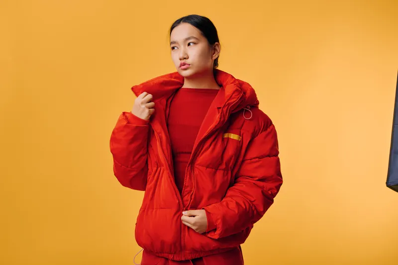 Young woman wearing red winter jacket and red clothing standing against yellow background. Fashion portrait with casual style, cold weather outfit, and thoughtful expression for seasonal trend.