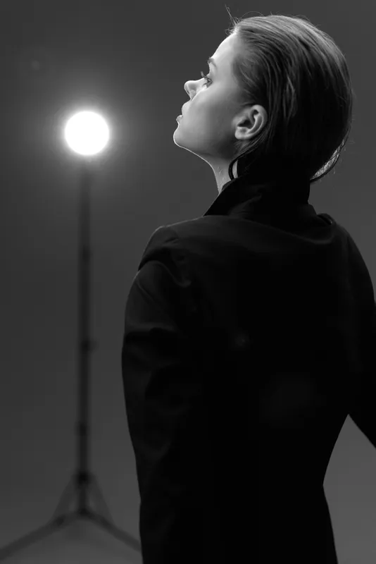 Woman in black outfit looking upward near bright light in black and white artistic photo.