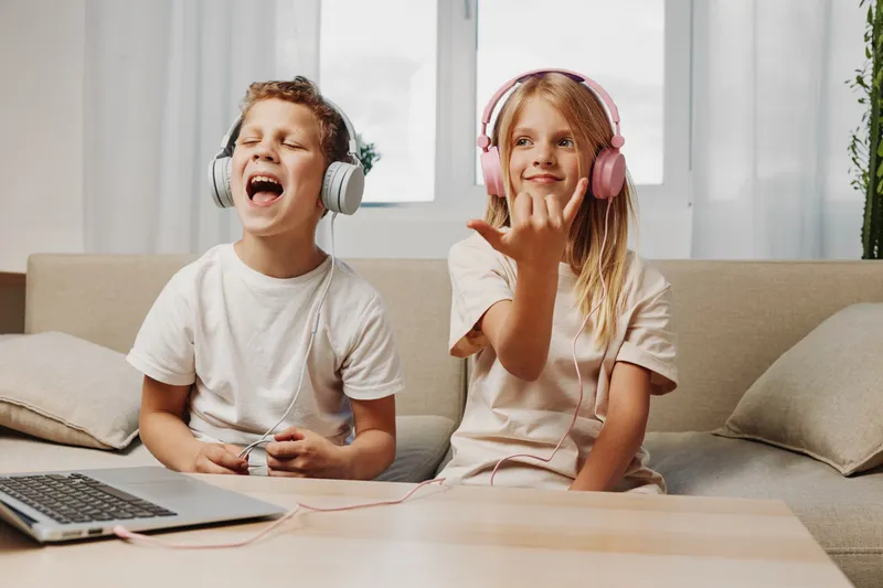 Two happy children wearing headphones sit on a couch enjoying music and games, expressing joy and entertainment in a bright living room setting with casual clothes.