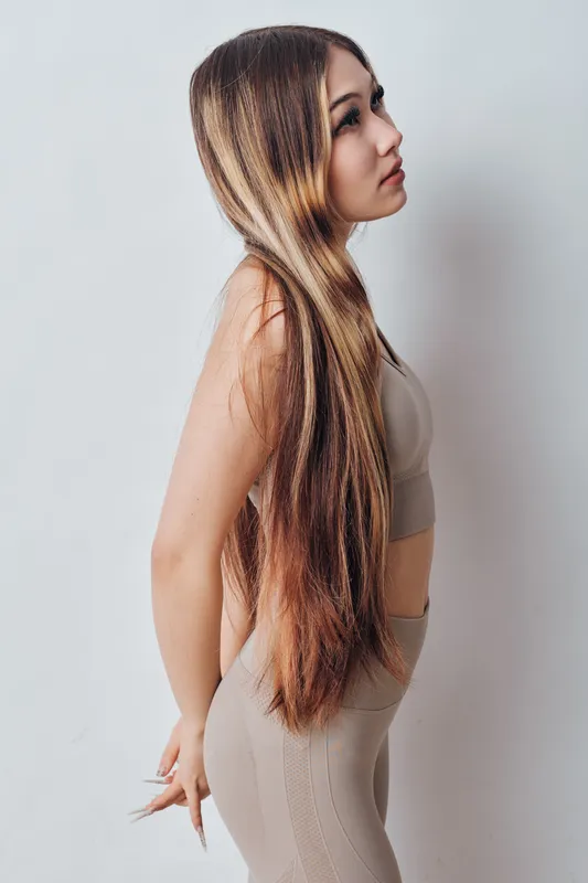 Young woman with long crimped hair standing sideways against white wall, wearing beige sleeveless top and pants, posing with relaxed hands behind back in casual style.