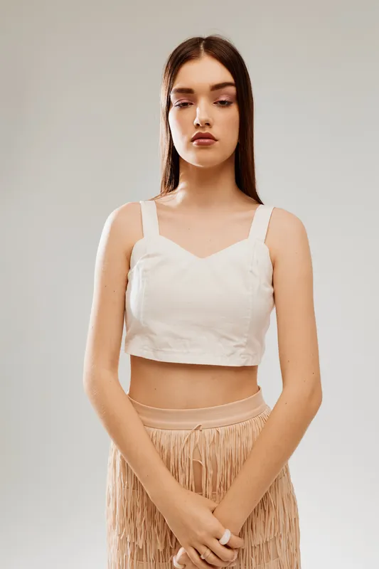 Young woman with long hair wearing a white crop top and beige fringe skirt stands with folded hands on a neutral background. Concept of woman beauty fashion and style.