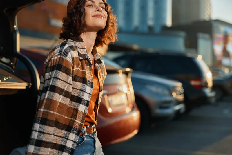 Woman leaning on car trunk in parking lot at golden hour glow, smile in plaid shirt and jeans with sunlight, creating candid lifestyle authenticity, mindful living and emotional storytelling vibe.