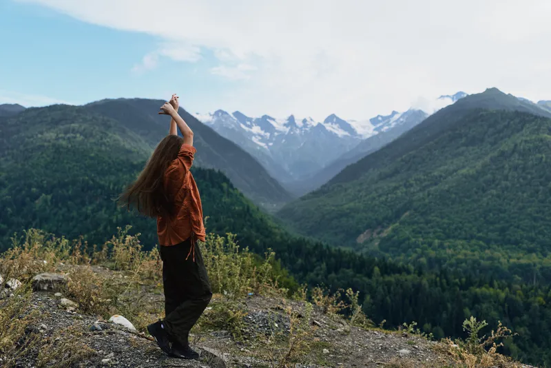 Person stands on rocky terrain amid a dramatic mountain landscape with arms raised, embodying hiking adventure and freedom as vast valleys and alpine peaks stretch into daylight