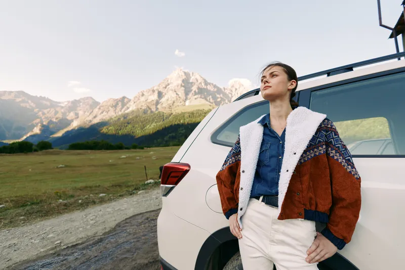 Woman leans against car at mountain roadside for travel portrait in outdoors, wearing warm jacket and denim shirt, road trip adventure with scenic nature, meadow view and confident pose.