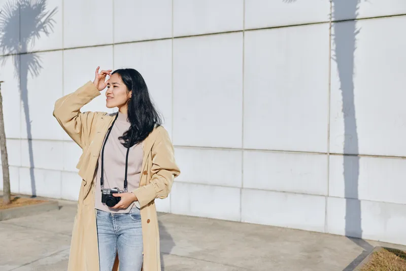 Woman shielding eyes from bright sun with her camera, outdoor scene, wearing a beige coat and jeans, casual street style moment captured in daylight