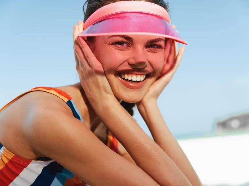 Joyful woman wearing a colorful striped swimsuit and pink visor smiles at the camera on a sunny beach, radiating carefree summer energy, bright mood, and vibrant outdoor vibe.