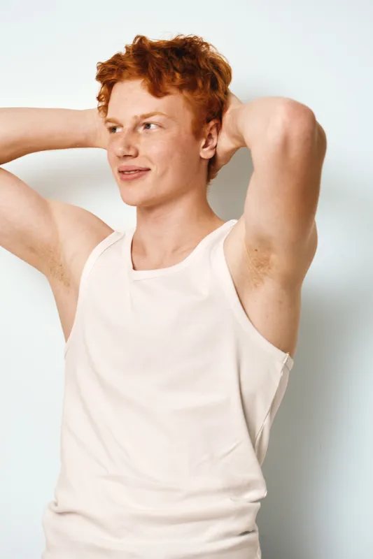Young man with red hair and fair skin wearing white tank top with arms raised looking away with thoughtful expression against white background.