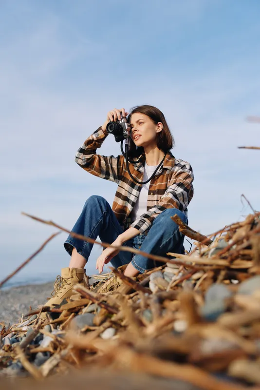 Woman with camera seated on rocky shore, exploring rugged coast in plaid shirt and jeans, capturing coastal scenery with a focused expression by the sea.