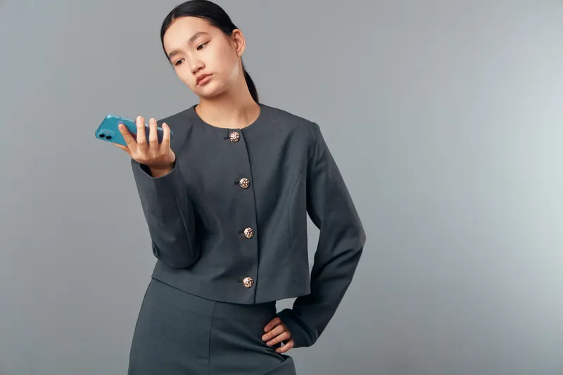 Woman in gray business suit holding smartphone with thoughtful expression against neutral background for technology, communication, and professional lifestyle concept.