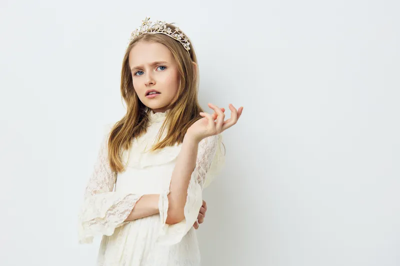 A young girl with a crown expresses confidence while wearing a white dress against a neutral background, embodying themes of royalty and elegance