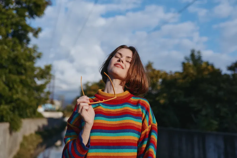 Young woman in a multicolor striped sweater holding sunglasses, smiling with eyes closed under a sunny sky on a suburban street, bob haircut, casual outfit, relaxed lifestyle moment.
