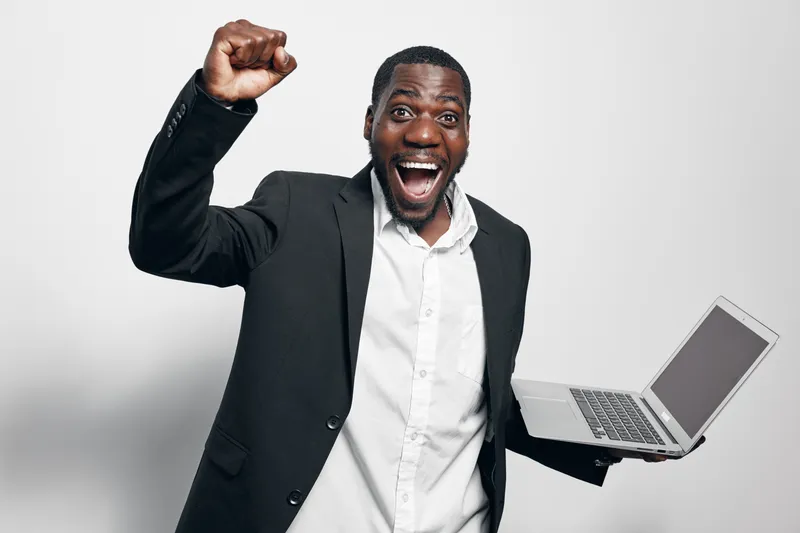 African man with dark skin in black suit holding laptop and celebrating success with raised fist and open mouth on white background. Business, technology and achievement concept.