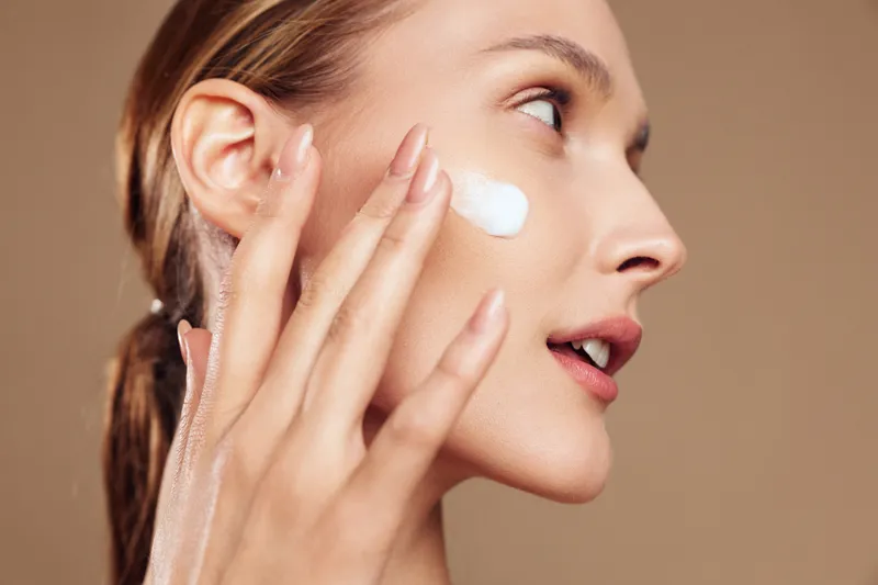 A young woman applies moisturizing cream to her cheek with fingers against a neutral background. The close-up highlights skincare, beauty, and facial care concepts.