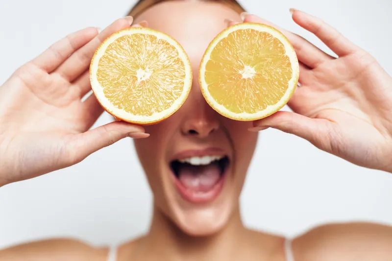 A cheerful young woman holds two halves of a fresh orange in front of her eyes, smiling widely with an open mouth. Concept for healthy lifestyle, freshness, and fun.
