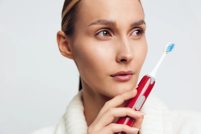 Young woman holding red electric toothbrush near face, thoughtful expression, clear skin, white background, personal hygiene concept, oral care routine, closeup portrait.