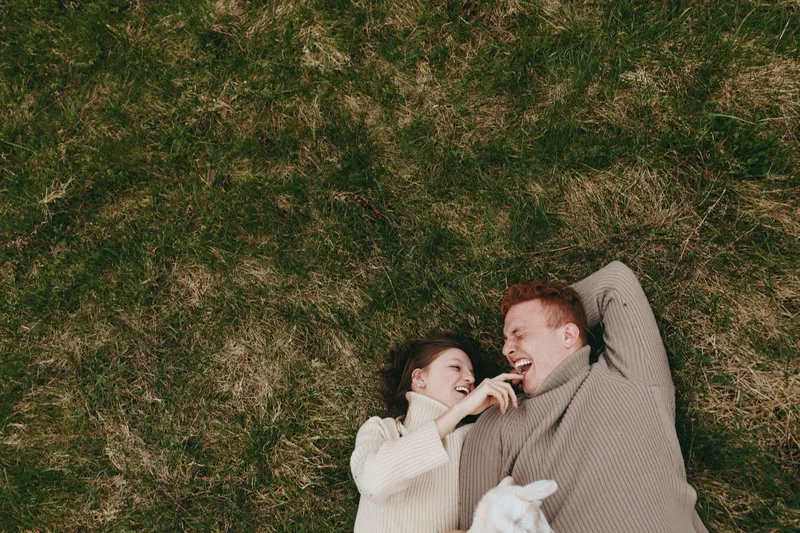 Casual couple outdoors, lying on grass, laughing together under sunlight, relaxed moment, gentle breeze, natural colors, goat kid nearby adds charm to a warm countryside scene