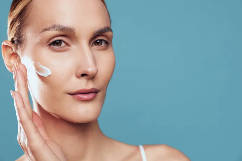 A close up of a young woman applying cream to her cheek, showcasing a skincare routine in a clean studio setting, conveying calm focus and self care.