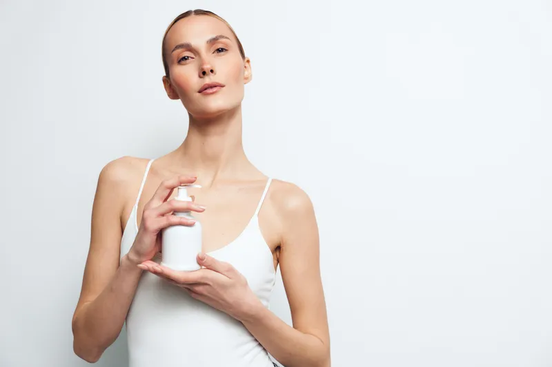 Young woman in white tank top holding a white bottle with pump dispenser. Minimalist studio background with soft lighting. Concept of skincare, beauty, health, and wellness.