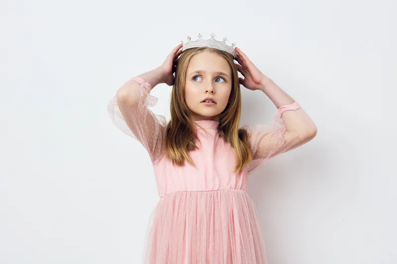A young girl in a pink tulle dress with a crown, looking surprised against a plain white background, evoking feelings of fantasy and innocence