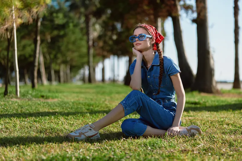 lifestyle woman in modern boho-western denim street style sitting on grass with sunglasses and red bandana, film color effect, relaxed outdoor summer mood