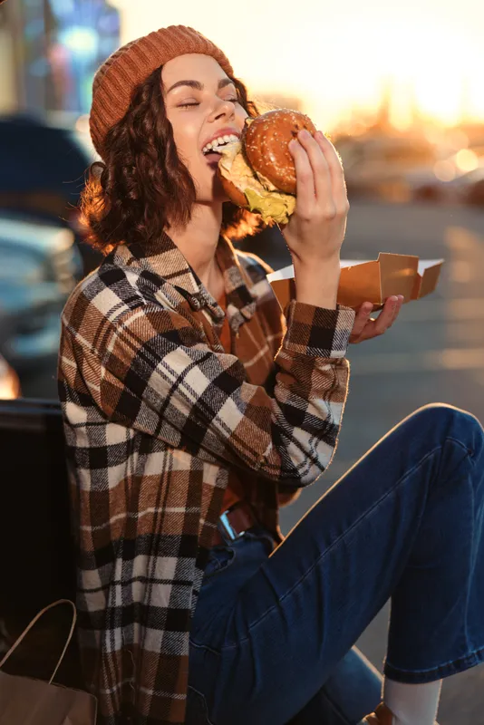 Woman eating burger outdoor at sunset with a joyful smile, candid moment capturing authenticity and candid lifestyle. Golden hour glow, mindful living and emotional storytelling vibe.