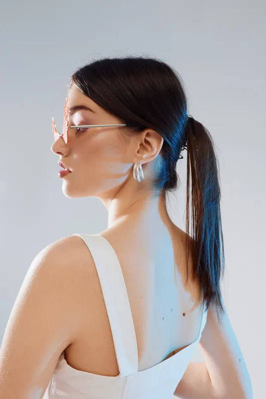 Young woman beauty fashion model with ponytail and glasses poses in white dress against light background. Studio portrait with soft lighting highlighting skin and style.