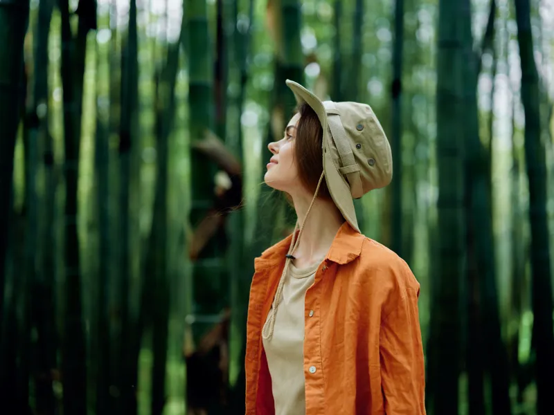 Woman in hat and orange jacket stands in a dense bamboo forest, portrait-style travel scene with nature vibes, looking up thoughtfully during outdoor exploration.