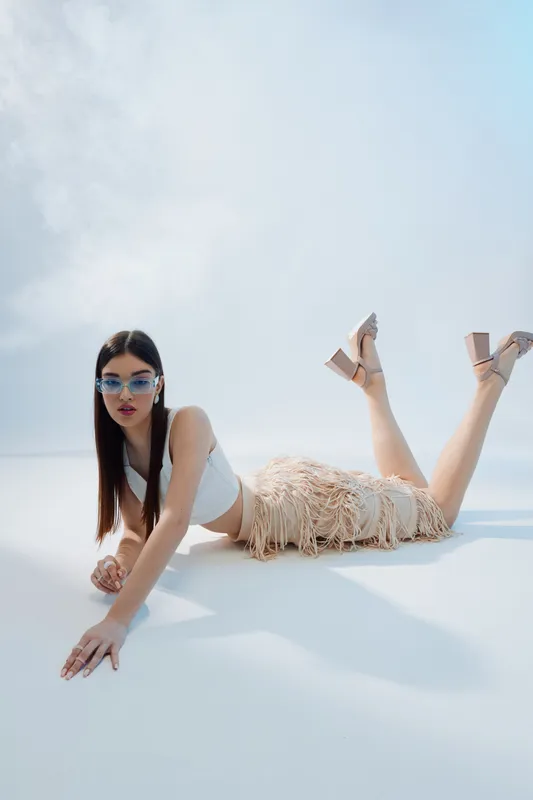 Young woman in stylish sunglasses and a fringed skirt lies on the floor against a light background. Concept of woman beauty fashion, summer style, and elegance.